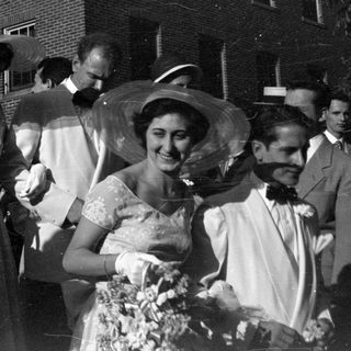 Group in formal attire and hats by brick building.