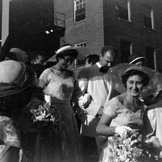 Group in formal attire and hats by brick building.