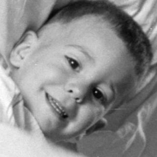 Young child lying in bed under striped blanket.