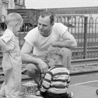 Man and children by railway tracks near industrial building.