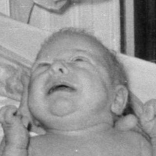 Woman in white uniform with crying infant in bassinet.