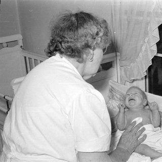 Woman in white uniform with crying infant in bassinet.