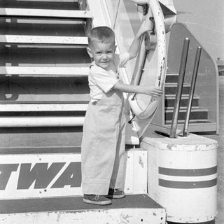 Child in overalls standing by TWA aircraft stairs.