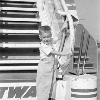 Child in overalls standing by TWA aircraft stairs.