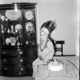 Child in headdress sitting by a birthday cake.
