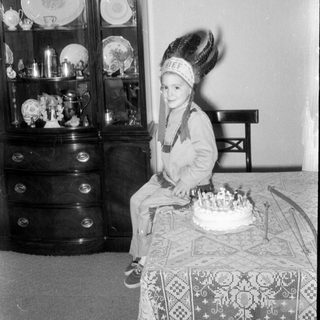 Child in headdress sitting by a birthday cake.