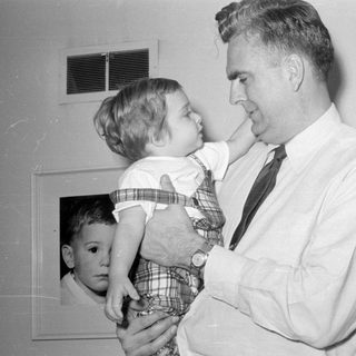Man in tie holding a child in plaid overalls.