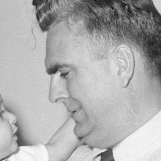 Man in tie holding a child in plaid overalls.