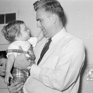 Man in tie holding a child in plaid overalls.
