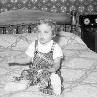 Child in plaid overalls sitting on a bed.