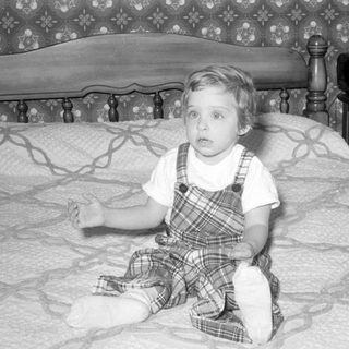 Child in plaid overalls sitting on a bed.