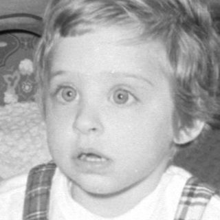 Child in plaid overalls sitting on a bed.