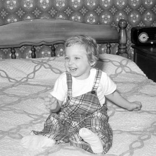 Child in plaid jumper sitting on a bed.