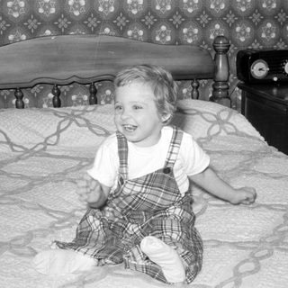 Child in plaid jumper sitting on a bed.