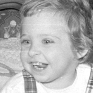 Child in plaid jumper sitting on a bed.