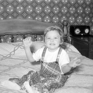 Child in plaid overalls sitting on a bed.