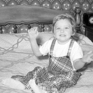 Child in plaid overalls sitting on a bed.