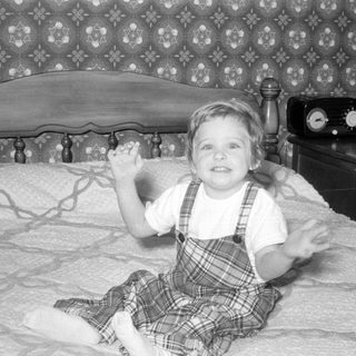 Child in plaid overalls sitting on a bed.