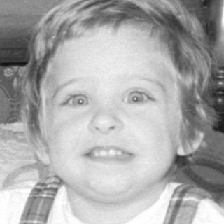 Child in plaid overalls sitting on a bed.