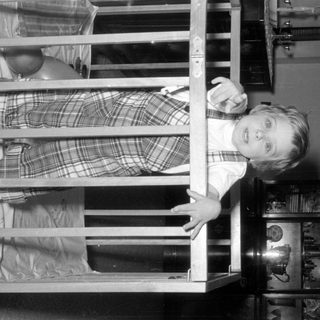 Child in plaid dress looking through crib bars.