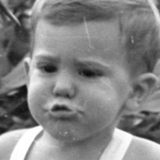 Toddler in white overalls walking on a lawn.