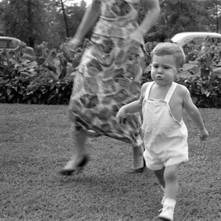 Toddler in white overalls walking on a lawn.