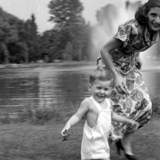 Child in white overalls running near a woman.