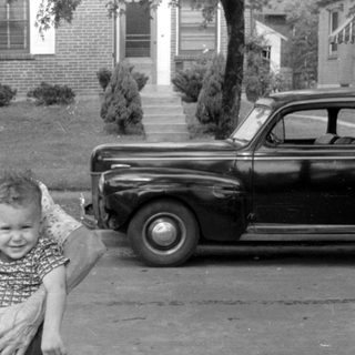 Woman holding a child in front of a vintage car.