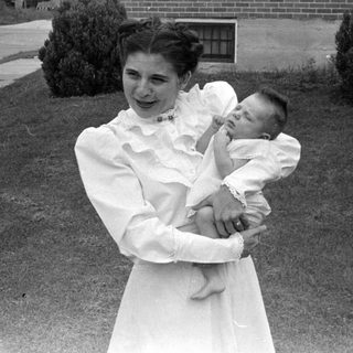 Woman in white dress holding an infant outdoors.