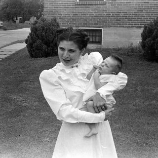 Woman in white dress holding an infant outdoors.