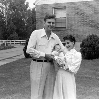 Man, woman, and infant in front of brick house.