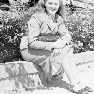 Woman in a suit sitting outdoors by foliage.