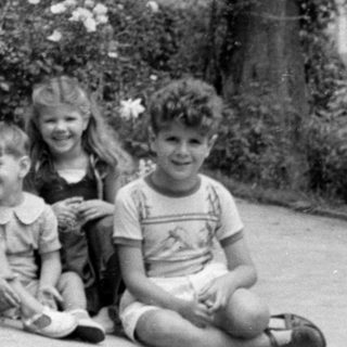 Six children sitting outdoors in front of bushes.
