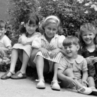 Six children sitting outdoors in front of bushes.