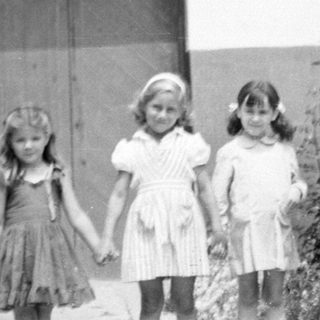 Three young girls standing before a building and shrubbery.