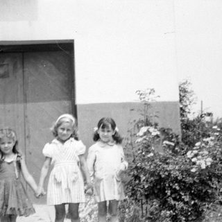 Three young girls standing before a building and shrubbery.