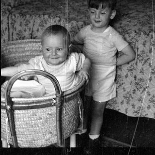 Infant in bassinet and toddler standing on bed.