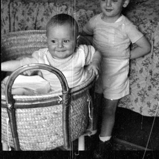 Infant in bassinet and toddler standing on bed.