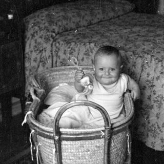 Infant sitting in a wicker bassinet indoors.