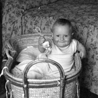 Infant sitting in a wicker bassinet indoors.