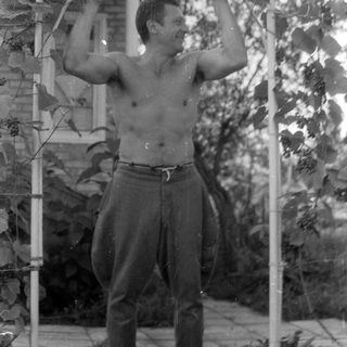Shirtless man standing under a garden trellis.
