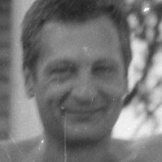 Shirtless man standing under a vine-covered trellis.