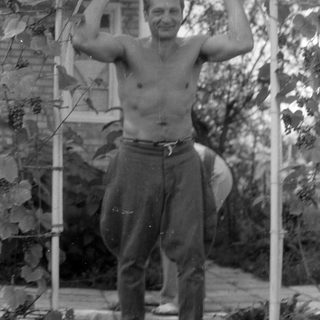 Shirtless man standing under a vine-covered trellis.