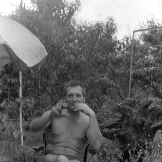 Shirtless man with a glass sitting outdoors.