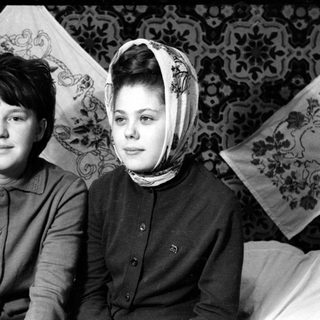 Two young women sitting before patterned wallpaper.