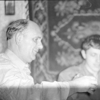 Two men indoors with a patterned wall hanging.
