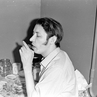 Man drinking from a glass at a cluttered table.