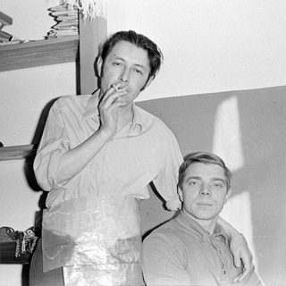Two men indoors, one wearing a plastic apron.